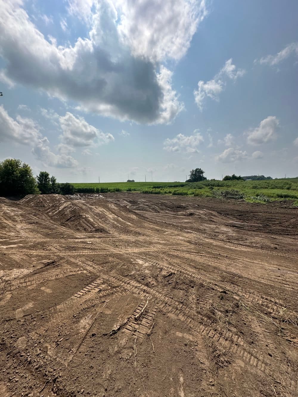 Cleared land with tire tracks, under a cloudy sky, near green fields and trees.