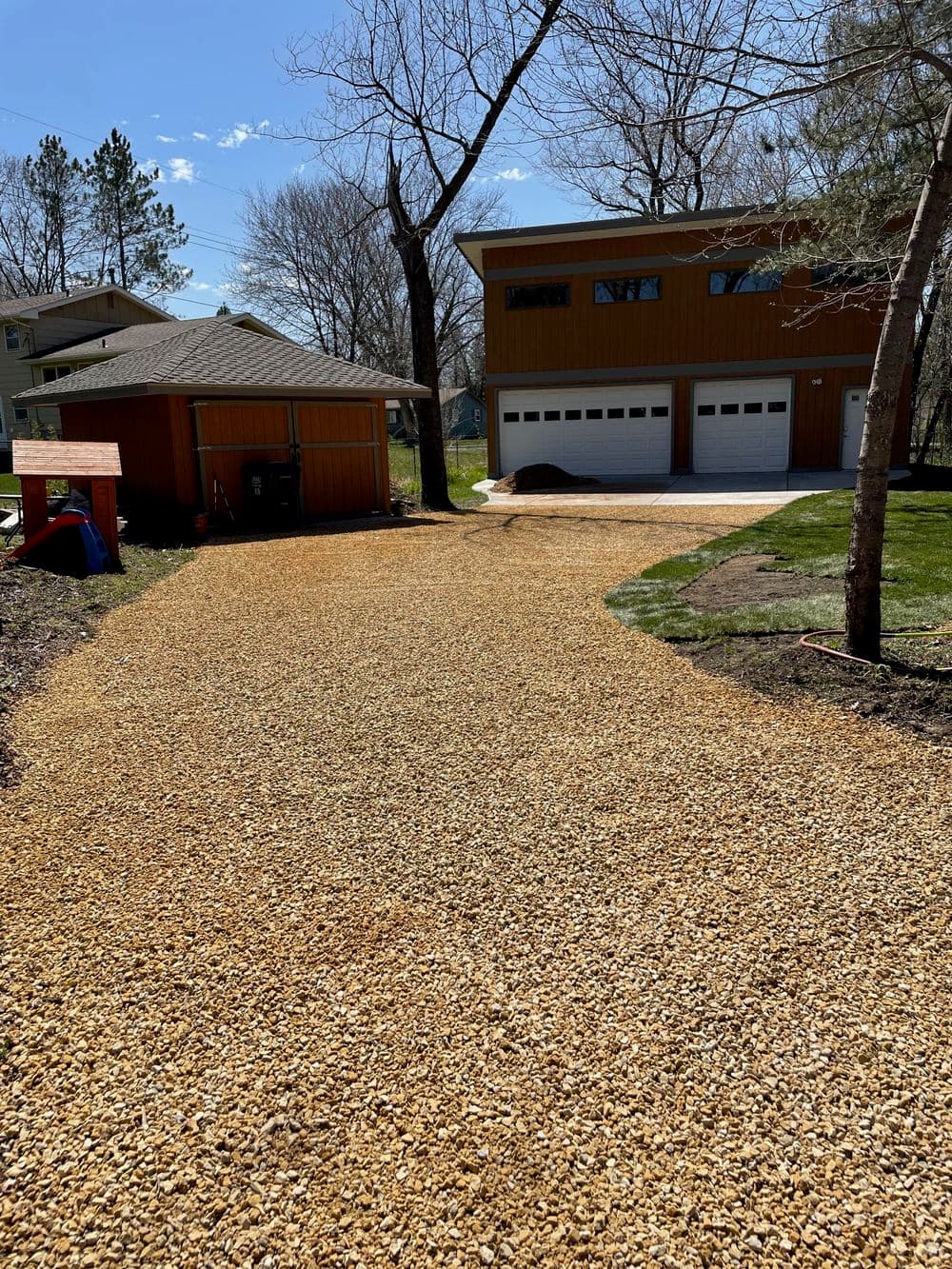 Gravel driveway leading to a modern house with two garages and trees in the background.