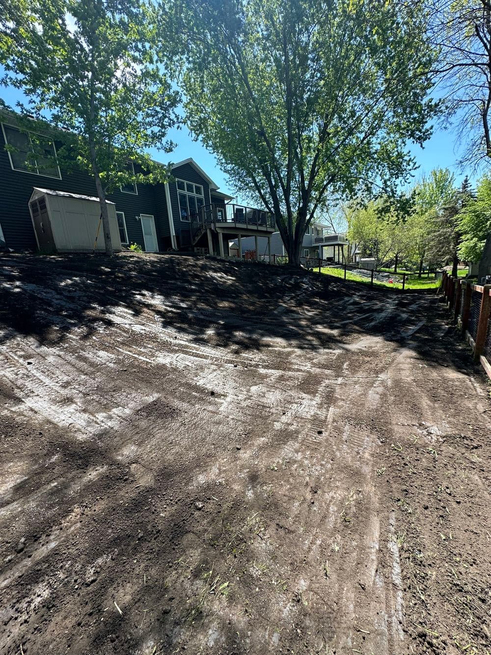 Landscape showing a sloped backyard with trees and a house in sunny weather.